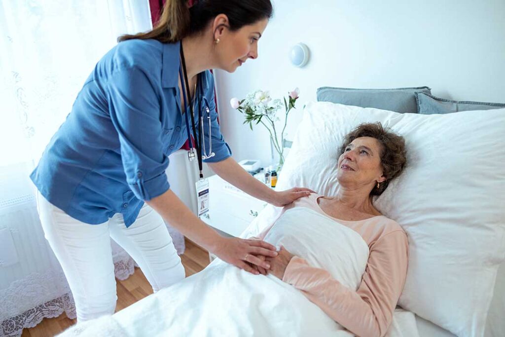 Two seniors and a caregiver share conversation at a dining table in a light-filled communal room.